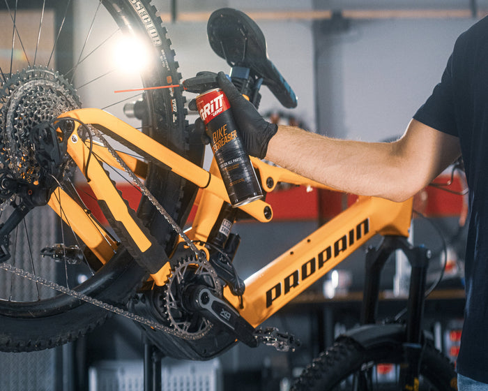 A person in a black t-shirt and gloves is applying a red spray canister to the orange Proton rear suspension of a mountain bike in a workshop setting.