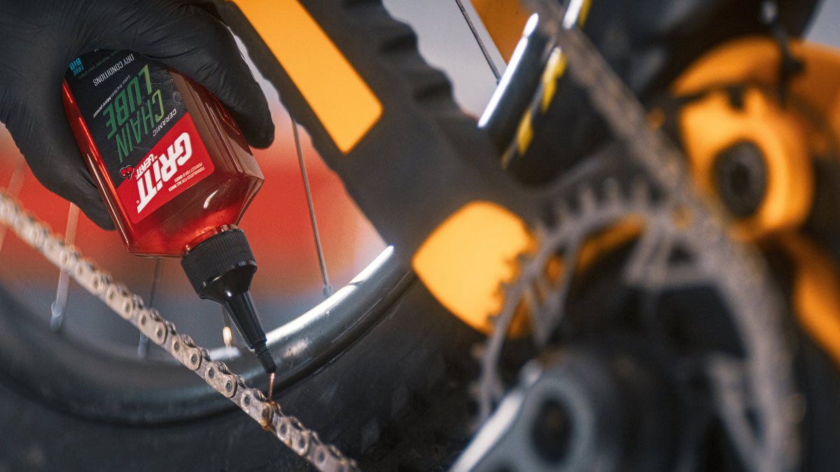 A close-up action shot of a person wearing a black glove applying grease from a red and black Gritt Lube 3 bottle to a bicycle chainring and chain. The background shows a blurred yellow and black mountain bike component.