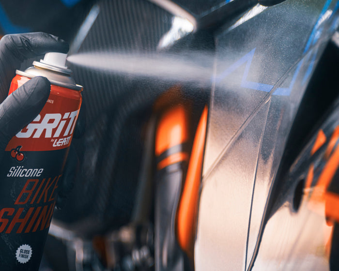A close-up action shot of a person wearing a black glove and spraying a can of Gritt Shine Silicone.