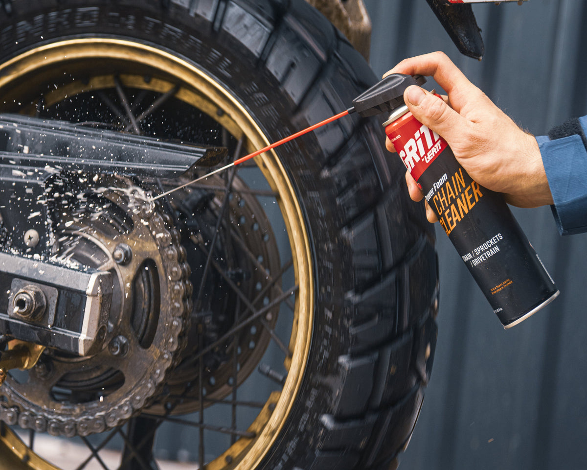A close-up action shot of a person using a foam cannon to spray motorcycle chain and chain guard cleaning foam onto a motorcycle's rear wheel and chain assembly. The person's hand and arm are visible on the right side, wearing a blue sleeve, against a dark gray background.
