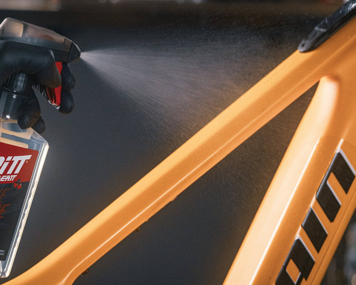 A person wearing a black glove sprays Grit Bike Shine onto a bright orange bicycle frame. The spray mist is visible, and the bottle of Grit Bike Shine is held in the foreground.