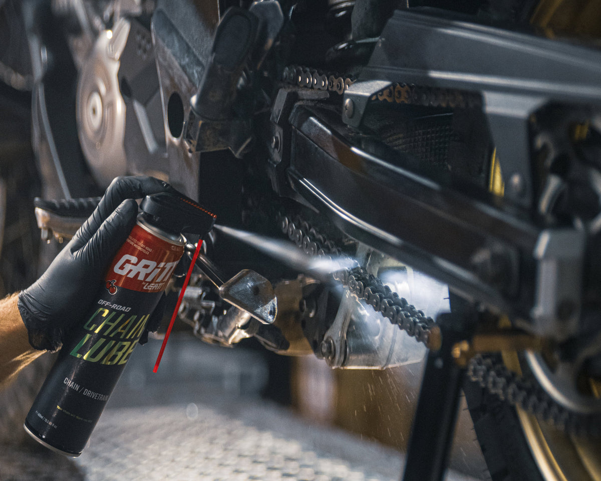 A man wearing safety glasses and a black t-shirt applies Grease Pro Chain Tensioner Grease from a black spray can to a motorcycle chain and sprocket in a workshop setting.