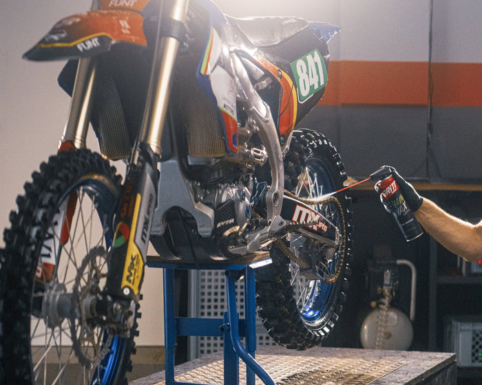 A man in a garage kneels beside a dirt bike on a lift stand, working on the motorcycle. The bike has orange, black, and white graphics with blue wheels.