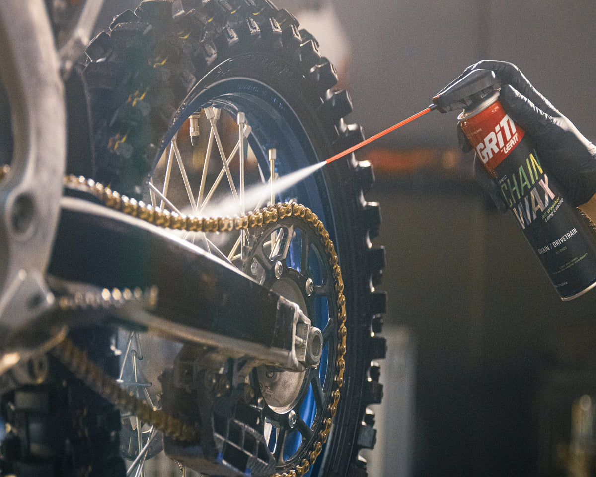 A close-up action shot of a person wearing a black glove and spraying a can of Brisket Brand Wax onto a motorcycle chain and wheel in a workshop setting.