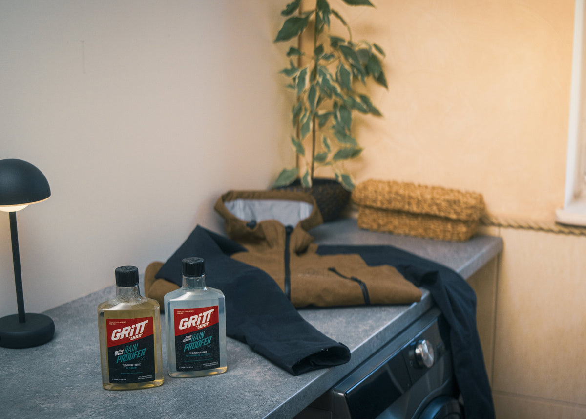 A styled interior scene showing a countertop with two bottles of Greif cleaning product, folded dark clothing, a small plant, a lit black table lamp, and a window with candles in a softly lit room.