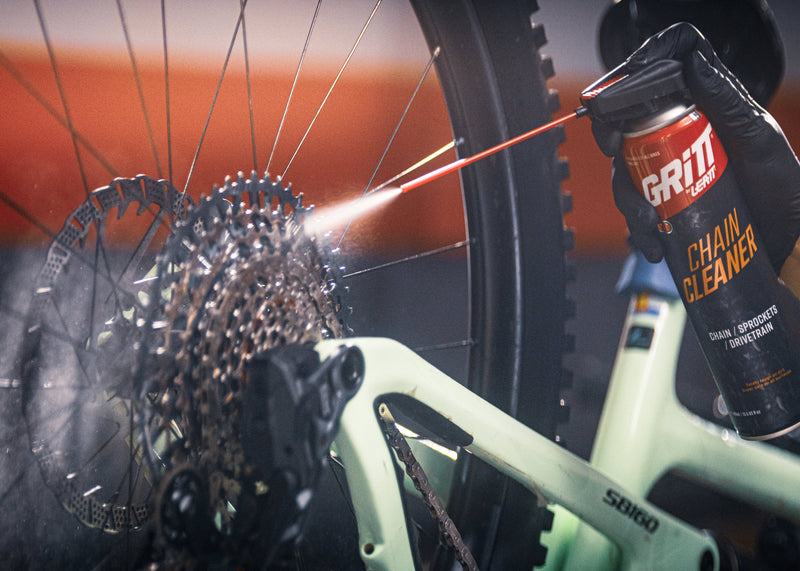 A person wearing a black glove sprays GRITT Brake Cleaner onto a bicycle cassette and rear wheel in a workshop setting.