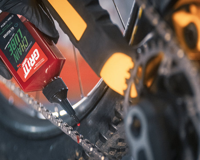 A close-up action shot of a hand in a black glove holding a red and black  bicycle chain oil bottle, applying oil to a bicycle chain near the rear cogset