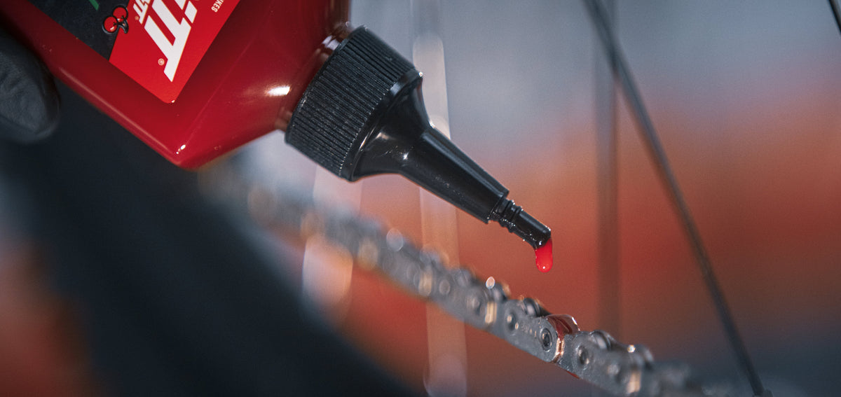 A close-up of a red bottle with a black applicator tip dispensing a small red liquid onto a bicycle chain, with a blurred background.