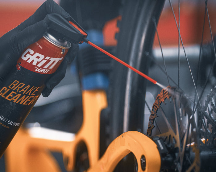 A close-up of a hand wearing a black glove spraying brake cleaner from a can labeled GRITT onto a bicycle disc brake rotor.