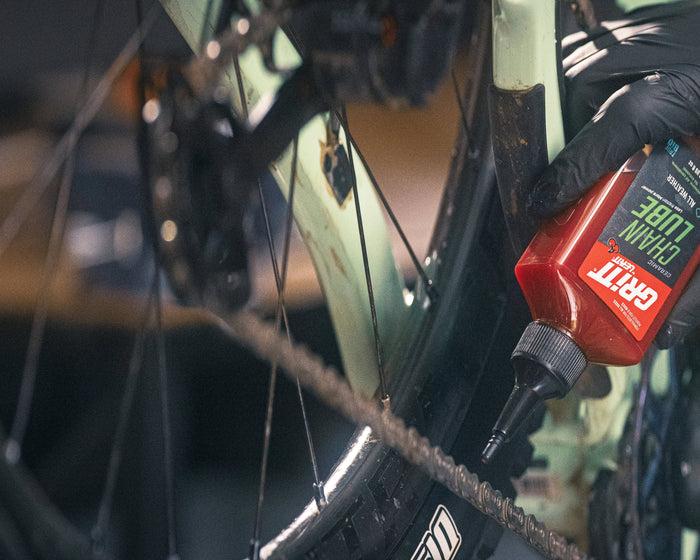 A close-up of a person wearing a black glove applying chain lube to a bicycle chainring.