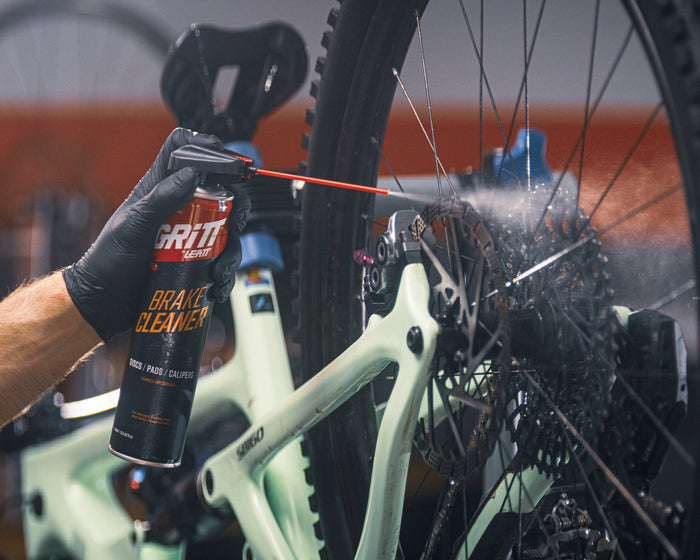 A person wearing a black glove sprays Brift Bike Chain Clean onto a bicycle's rear cassette in a workshop setting.