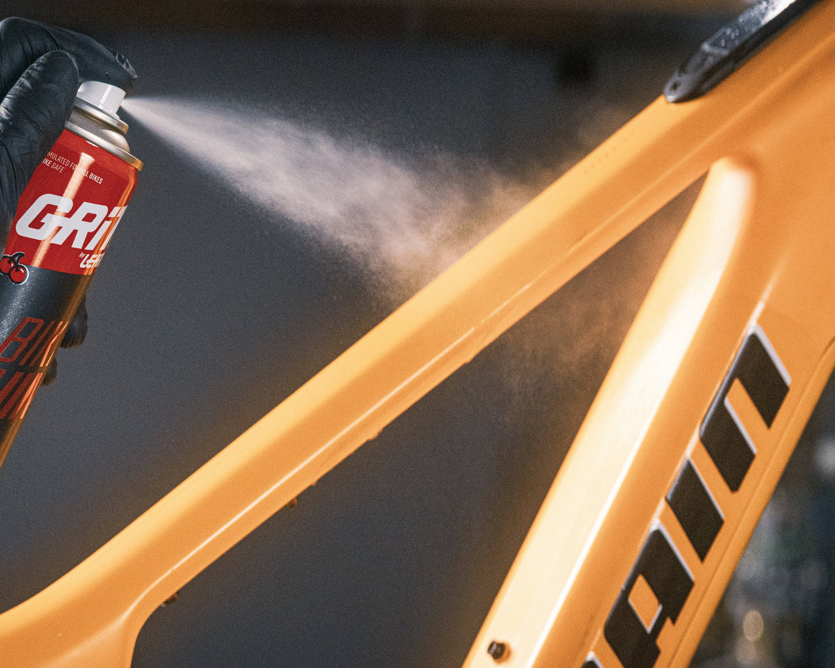 A close-up of a person's gloved hand holding a can of GRITT bike shine and spraying it onto an orange bicycle frame.
