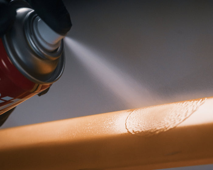 A close-up action shot of a black-gloved hand holding a red spray can and applying a lubricating mist to a textured orange surface.