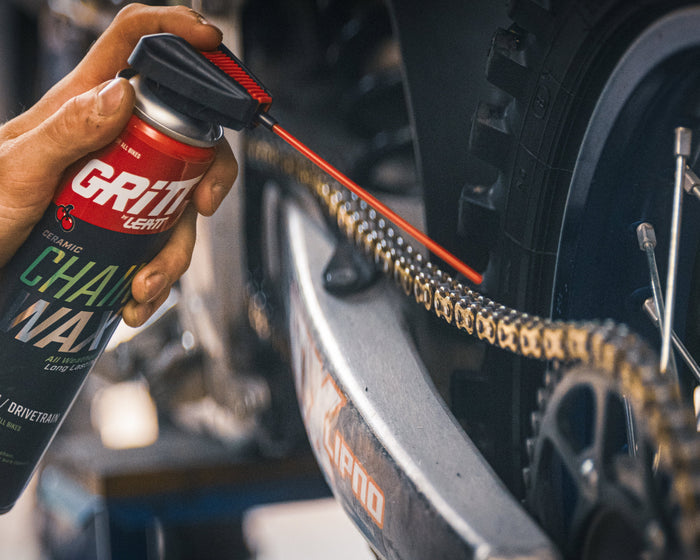A hand holding a can of Grit Chain Wax lubricant and spraying it onto a bicycle chain in an outdoor setting.