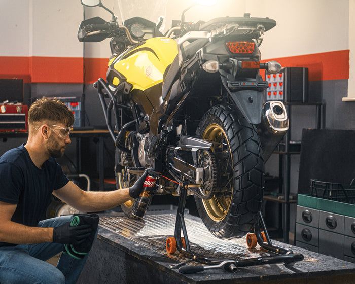 A man in a workshop kneels beside a yellow and black motorcycle on a lift platform, working on the bike. The garage has tools, shelves, helmets, and equipment visible in the background.