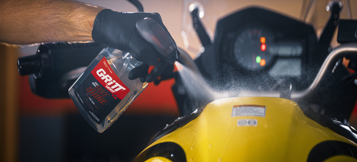 A person wearing a black glove sprays a clear bottle of GR-IT motorcycle bike shine fluid onto a yellow motorcycle in a garage setting.