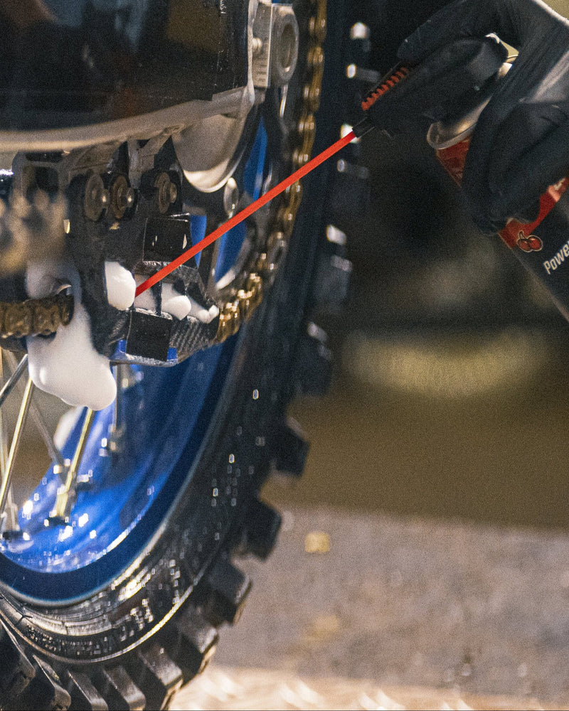 A close-up action shot of a person wearing a black glove spraying a can of chain lube onto a motorcycle's rear wheel and chain drive. The motorcycle is on a blue lift stand in a workshop setting.